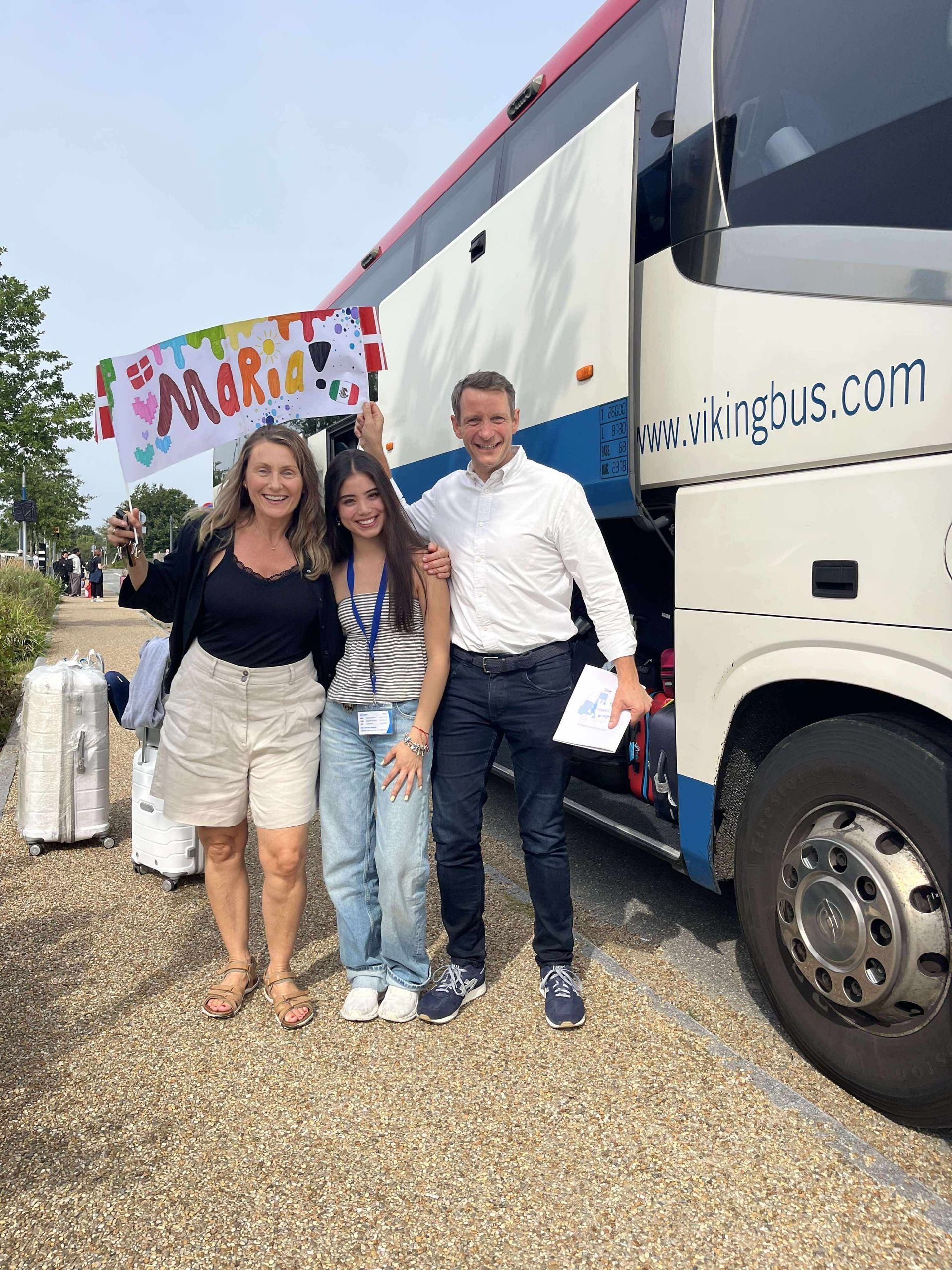 Tre personer poserer ved en bus, den ene holder et 