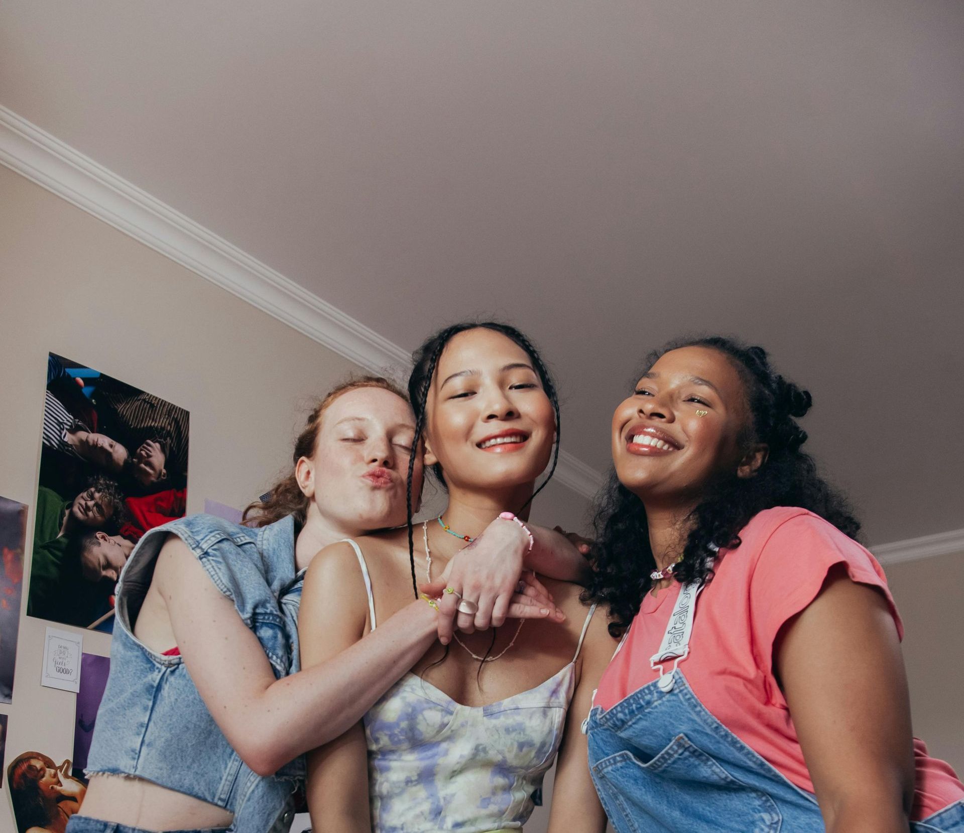 Tre smilende venner, arm om hinanden, poserer i et soveværelse. Den ene rynker læberne.