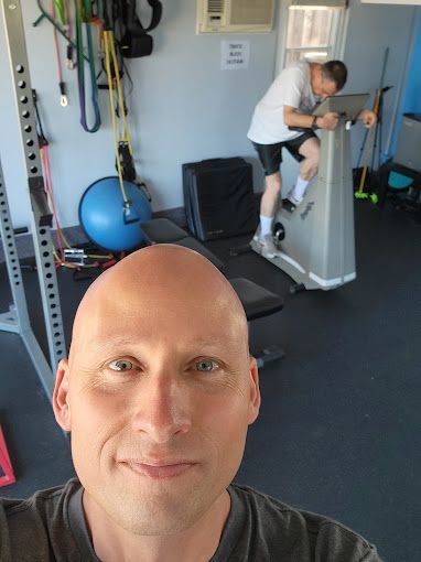 A bald man is taking a selfie in a gym while another man rides an exercise bike.