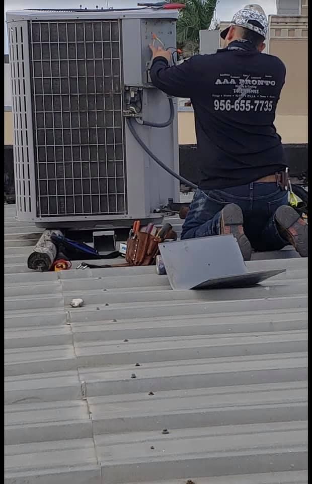 A man is kneeling on a roof working on an air conditioner.