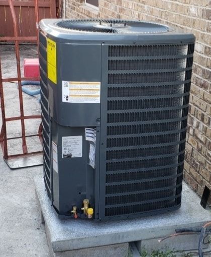 An air conditioner is sitting on top of a concrete platform in front of a brick building.