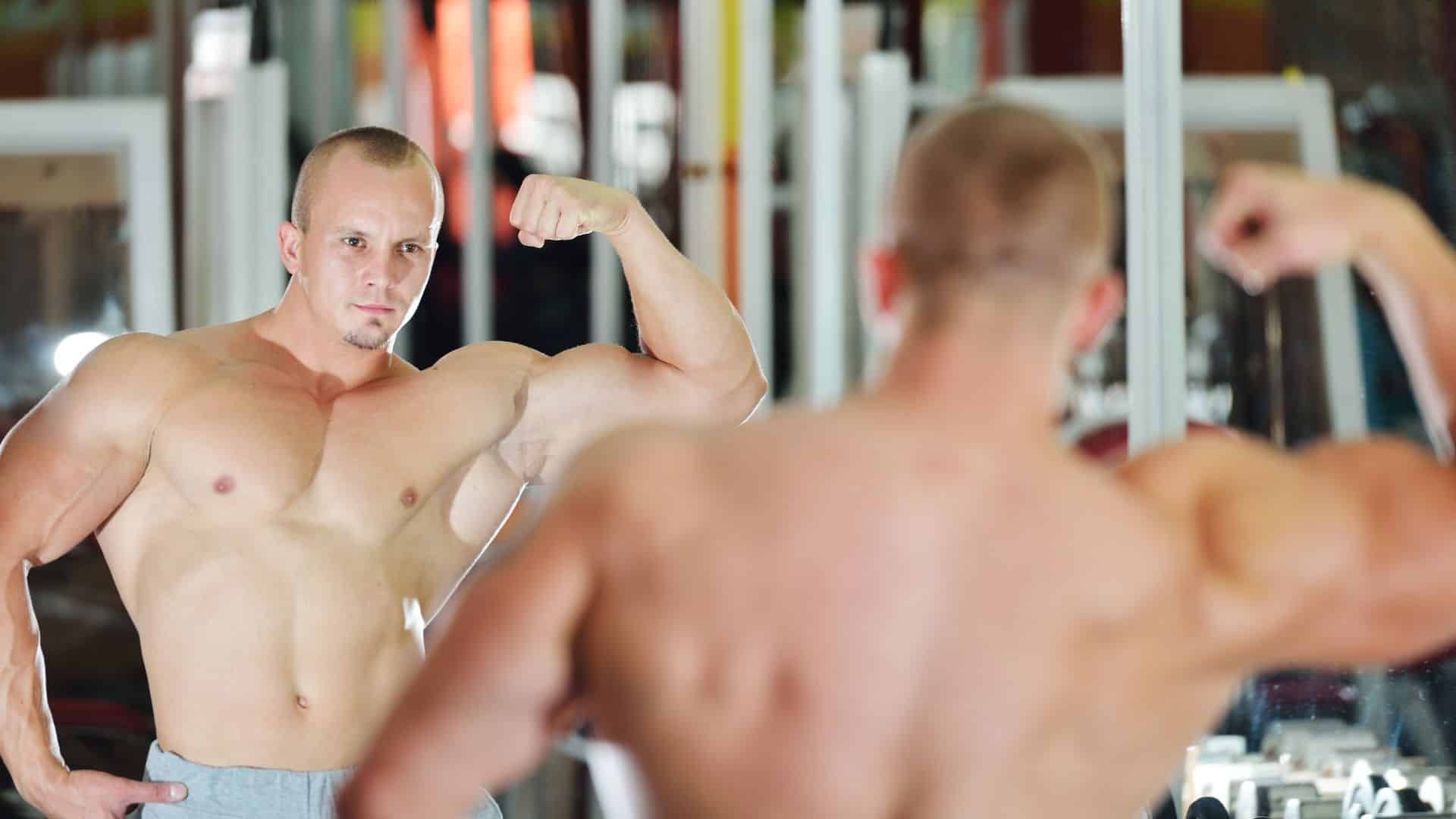 A shirtless man is flexing his muscles in front of a mirror in a gym.