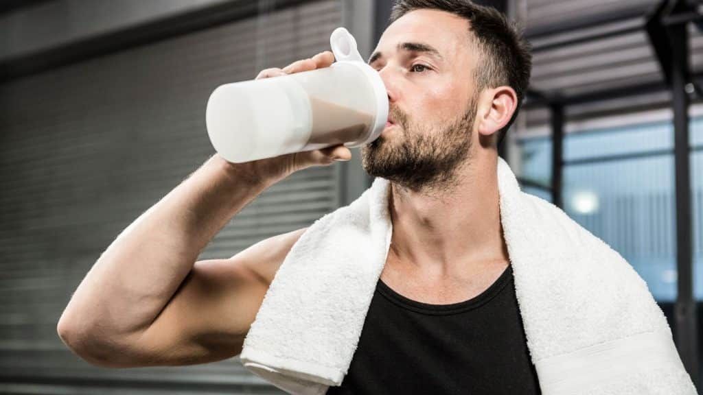 A man is drinking a protein shake from a shaker in a gym.