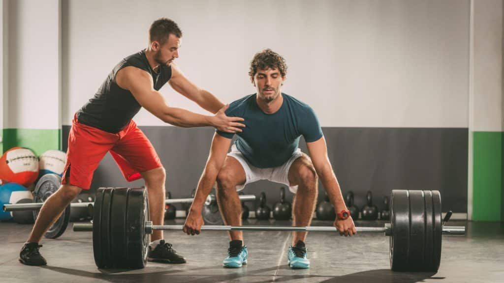 A man is helping another man lift a barbell in a gym.
