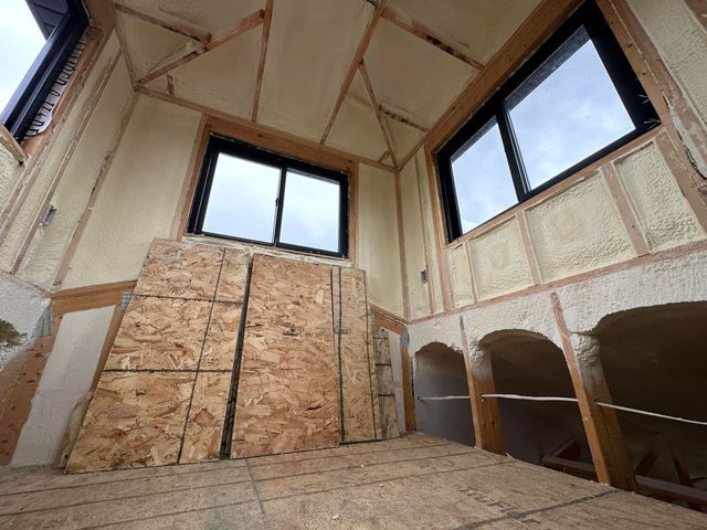 Interior of a small room under construction, insulated with spray foam. OSB panels lean against the wall. Two windows are visible.