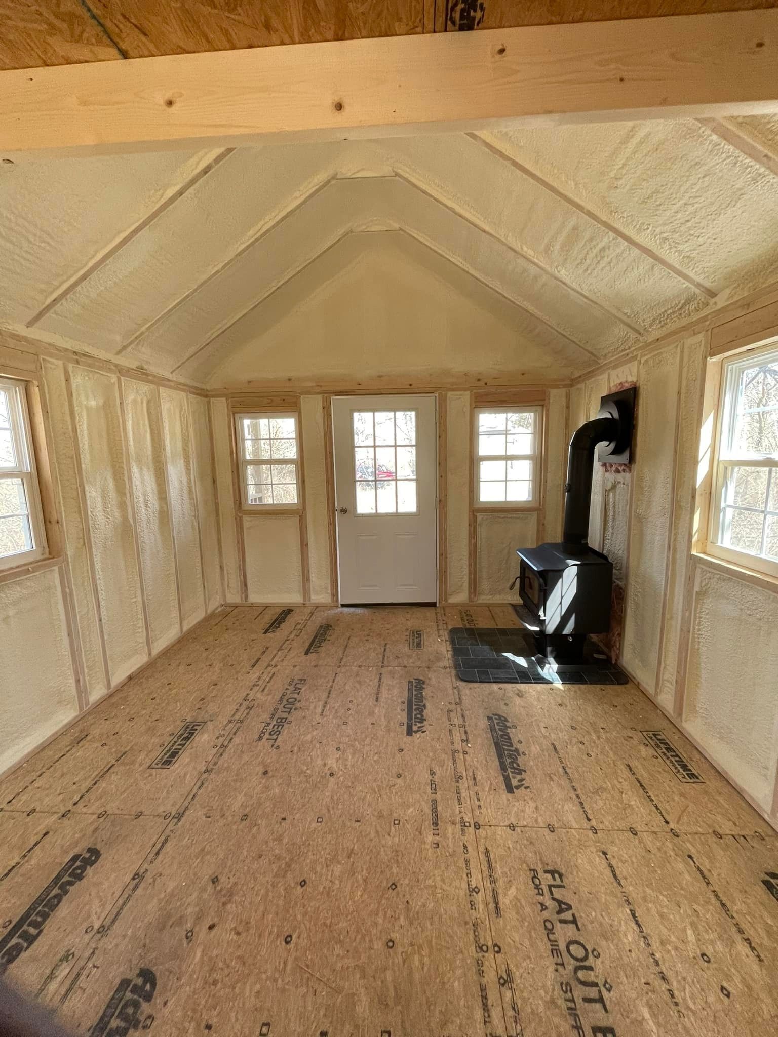 Interior of a small wooden cabin with spray foam insulation, wood stove, door, and windows.