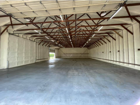 Interior view of an empty warehouse with spray foam insulation on the walls and ceiling, and a concrete floor.