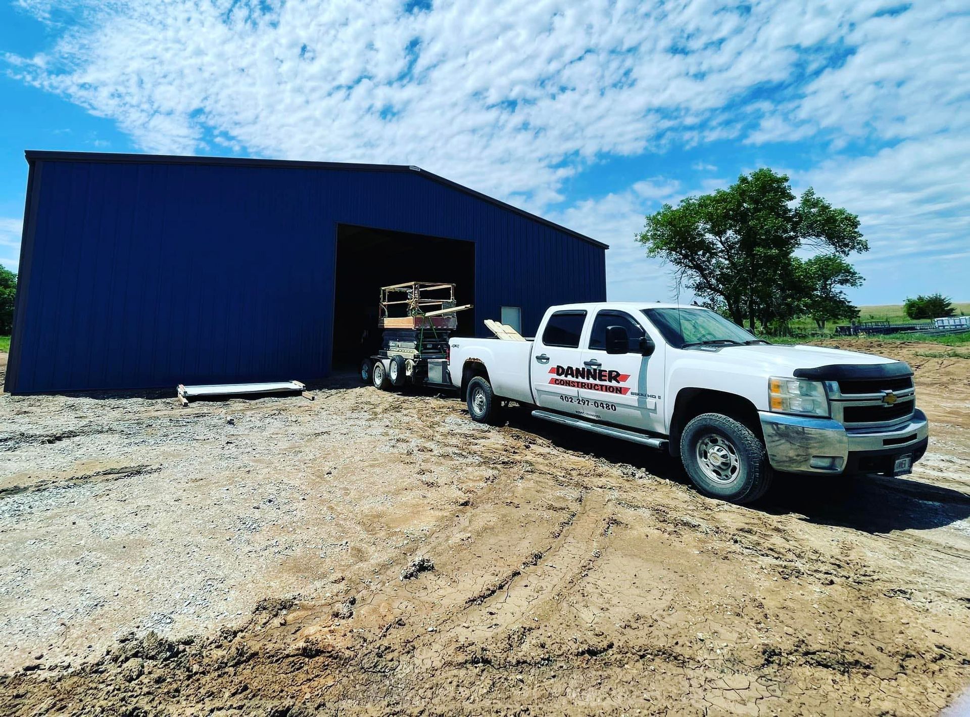 White truck towing a trailer with equipment outside a blue metal building under a bright sky.