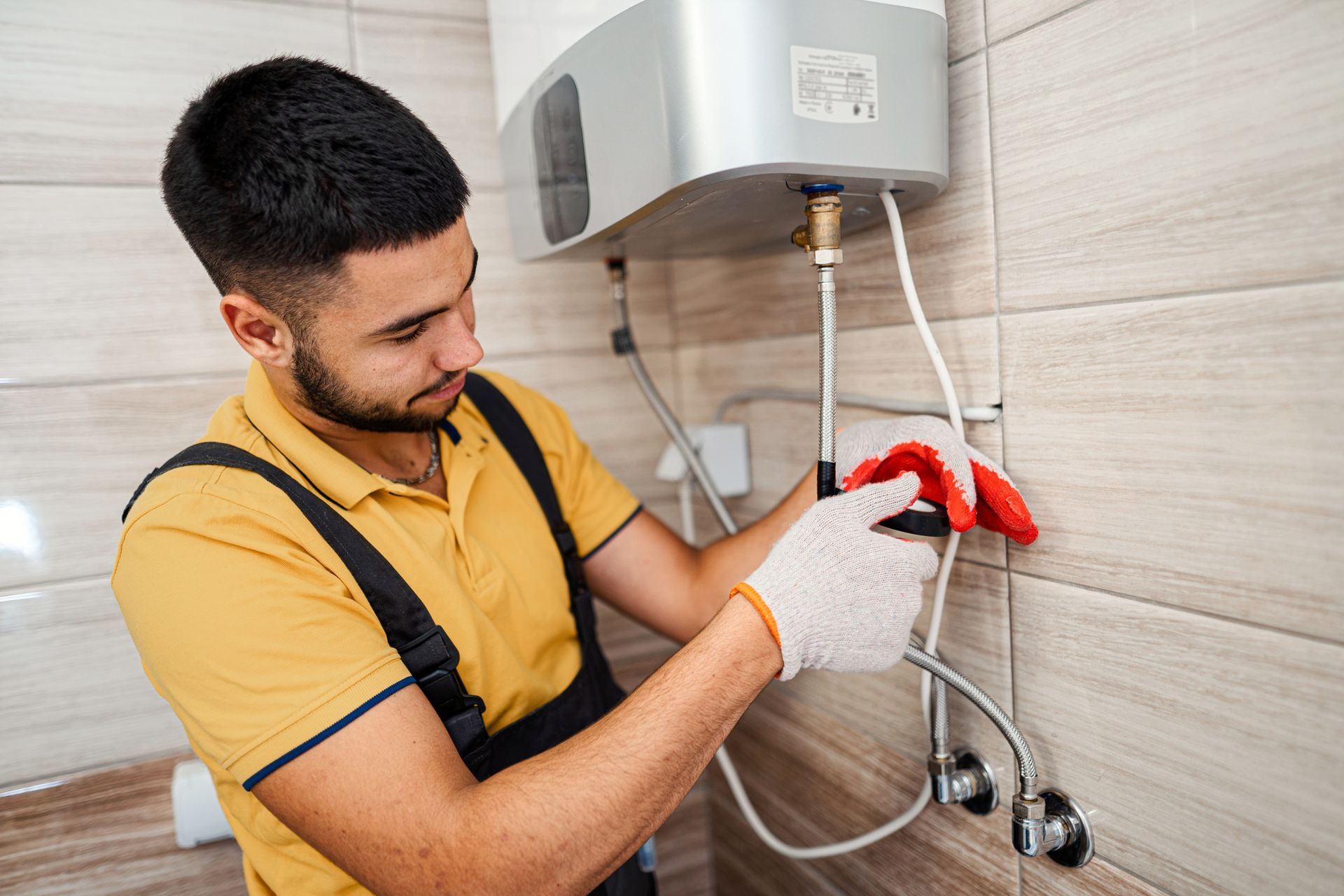 A plumber in a yellow shirt repairs a wall-mounted water heater with tools.