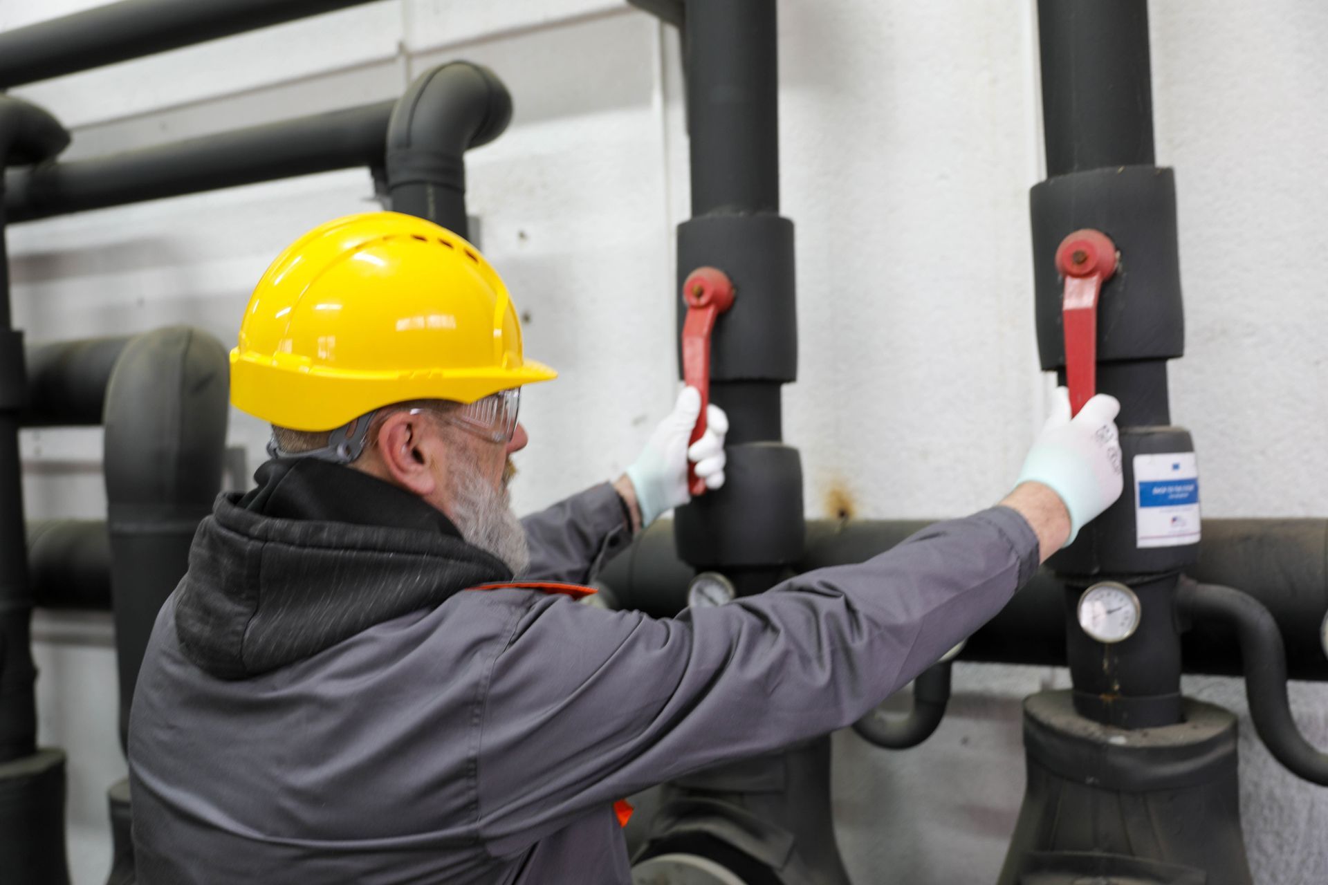A man in a hard hat & safety glass shuts off a valve on pipes in a commercial plumbing facility. A man in a hard hat & safety glass shuts off a valve on pipes in a commercial plumbing facility.