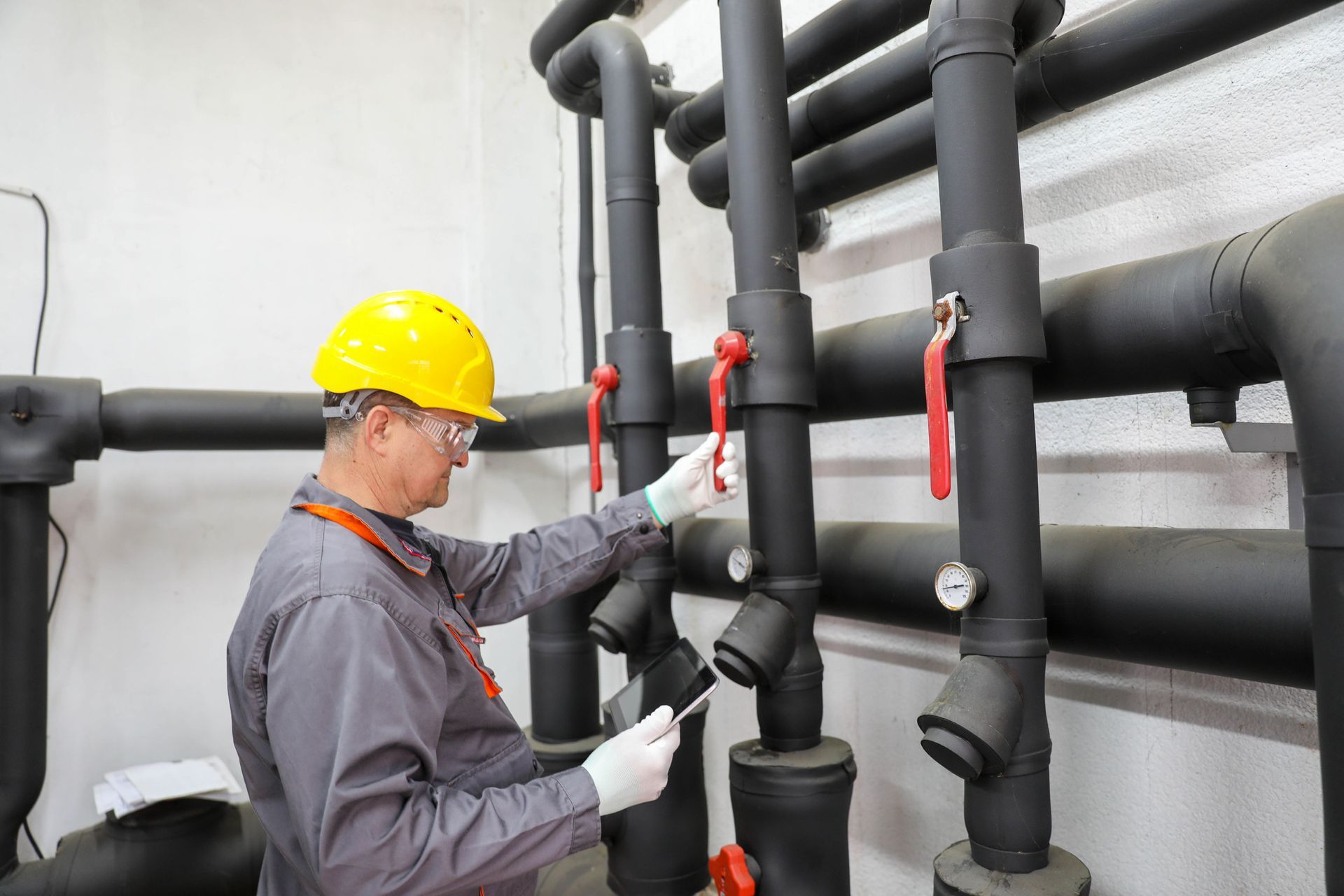 A man in a hard hat and safety glasses inspects pipes in a commercial plumbing facility. A man in a hard hat and safety glasses inspects pipes in a commercial plumbing facility.