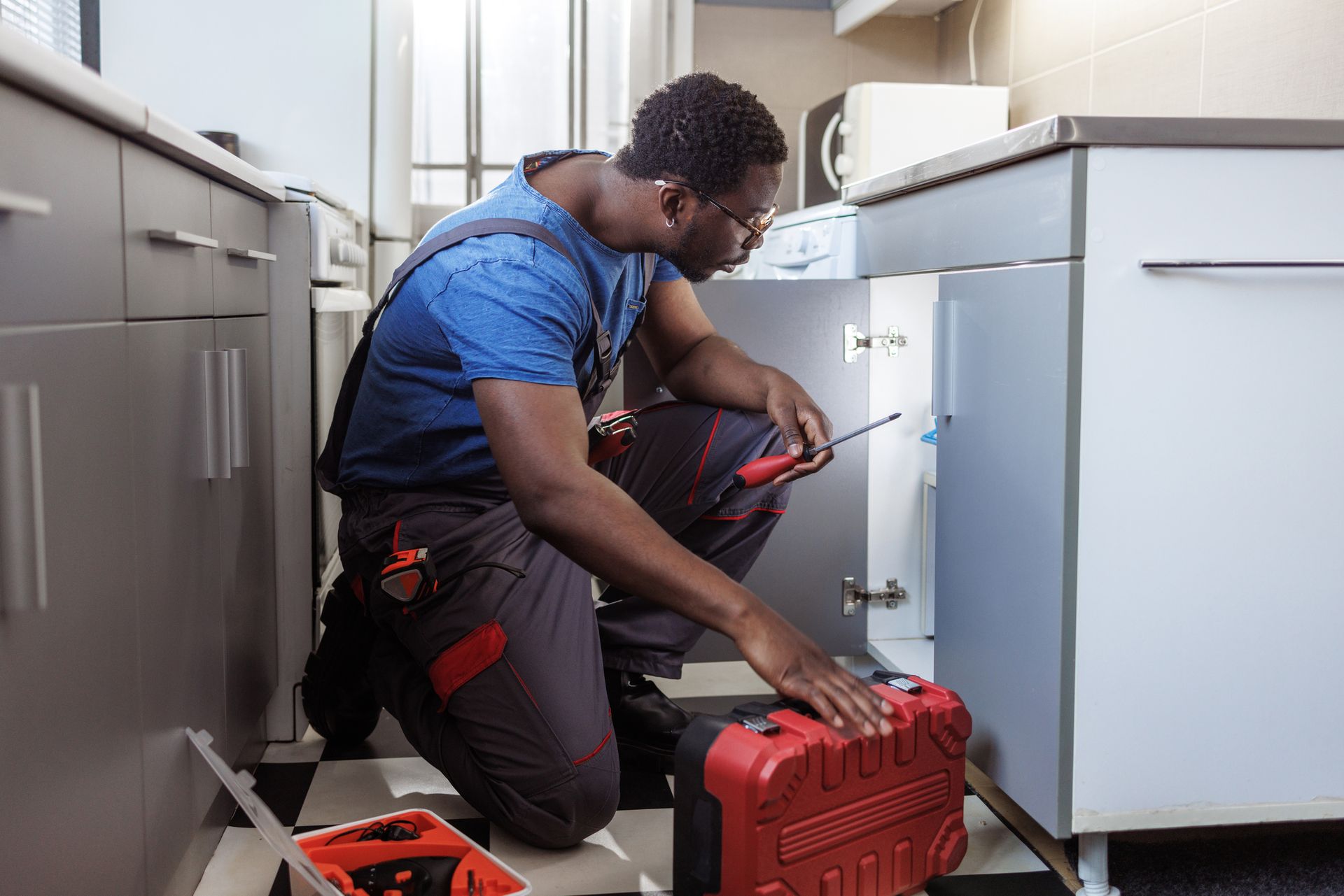 Skilled plumber offering plumbing services and using a wrench under kitchen sink with red tool case.