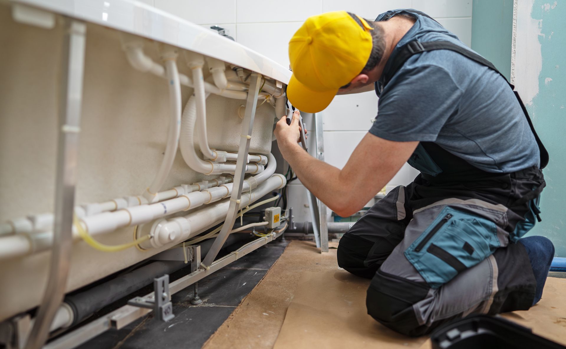 A plumber in a yellow cap is fixing pipes beneath a bathtub.