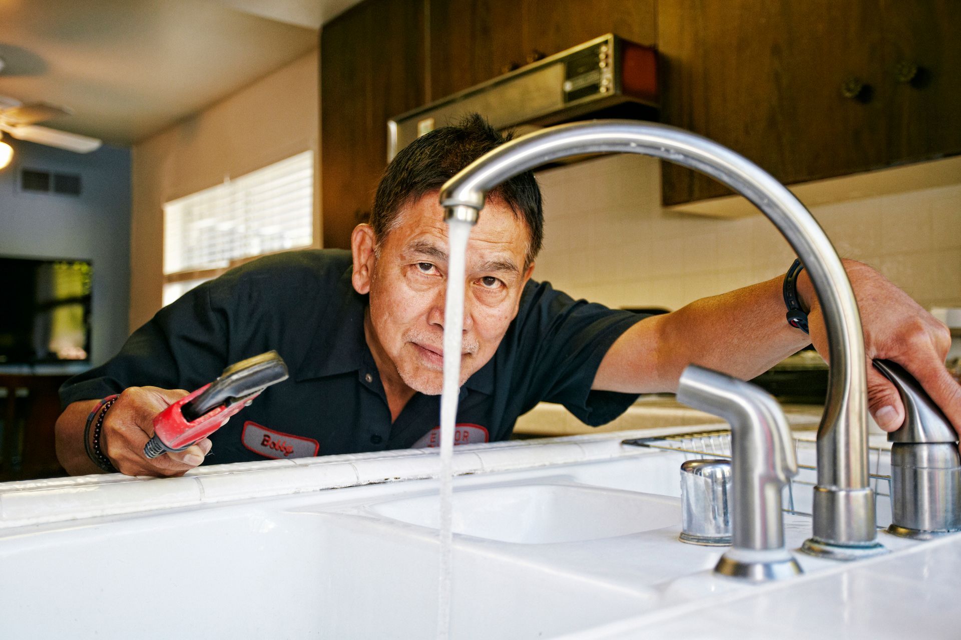 Plumbing repair technician inspecting kitchen faucet with water running and wrench in hand.