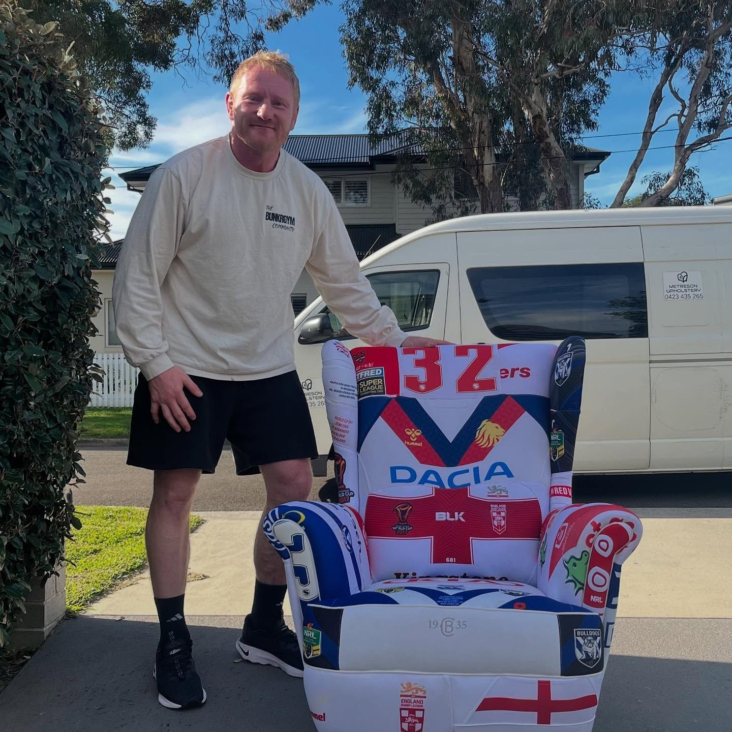 Man in shorts poses next to a decorated armchair with rugby symbols; white van in the background.— Metreson Upholstery In Unanderra, NSW