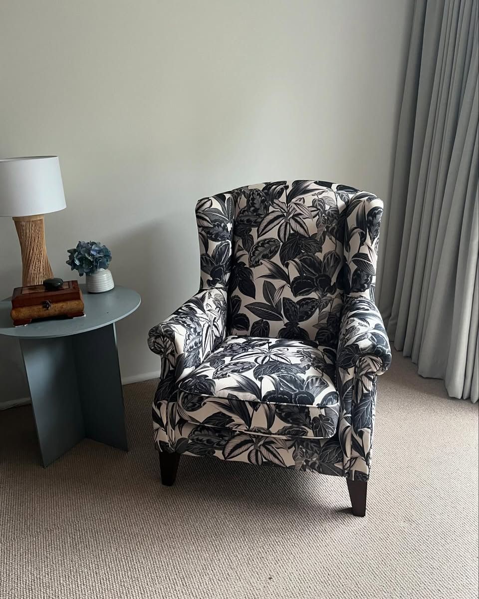 Wingback chair with floral pattern next to a side table with a lamp and vase, near a window with curtains.— Metreson Upholstery In Unanderra, NSW