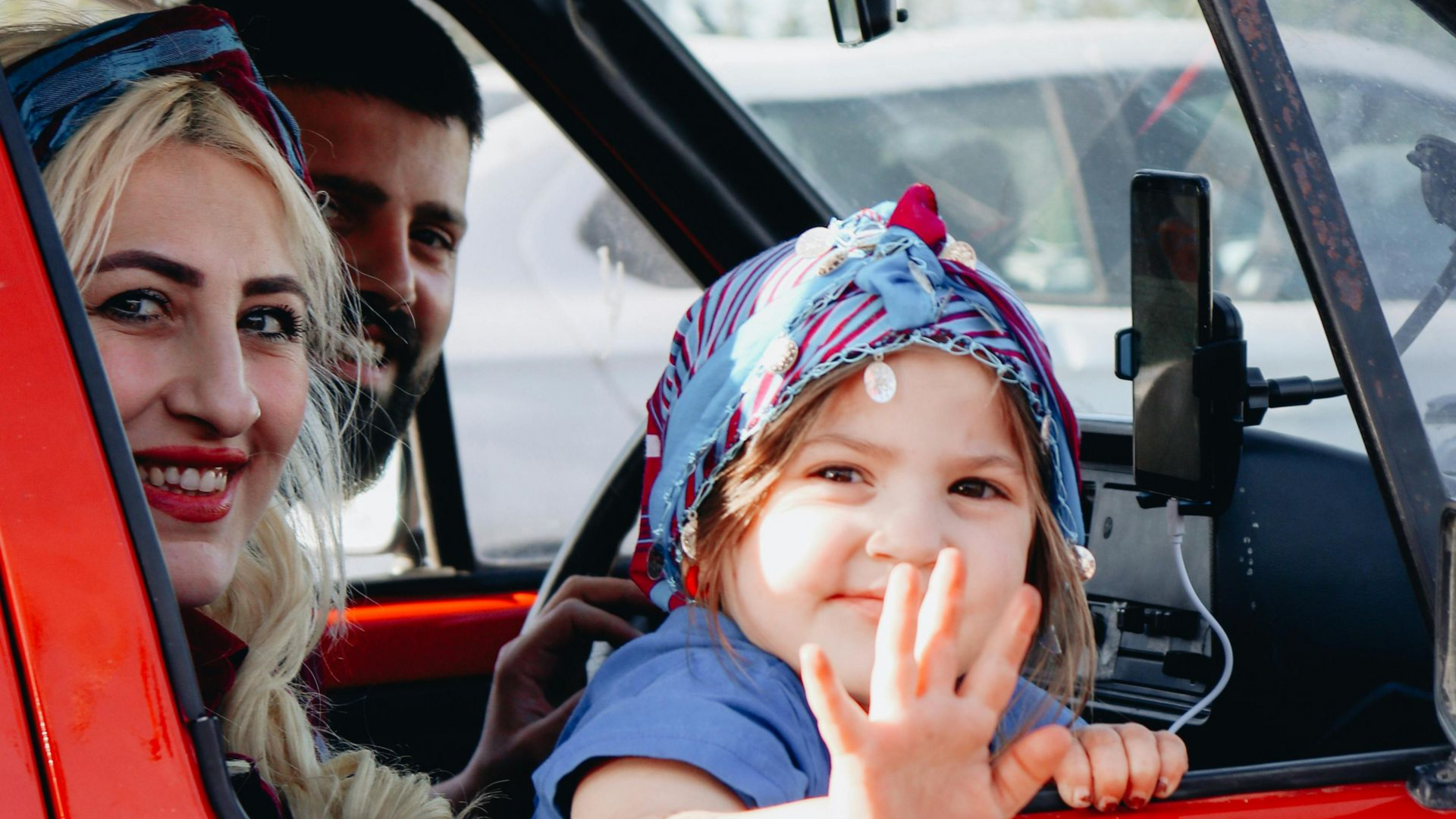 Family in a red car, child waving from window, smiling woman and man in background.