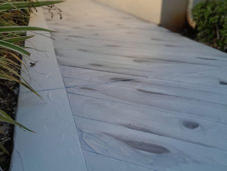 A concrete walkway with a wood grain texture is surrounded by plants.