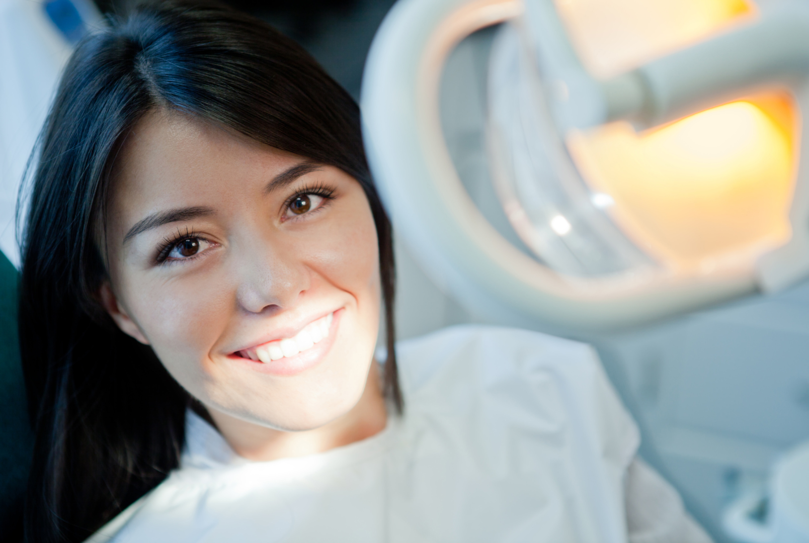 Woman smiling at dentist, lit by overhead lamp; dental office setting.