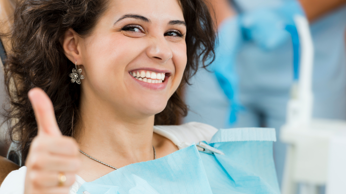 Woman in dentist chair smiles and gives thumbs-up, wearing bib.