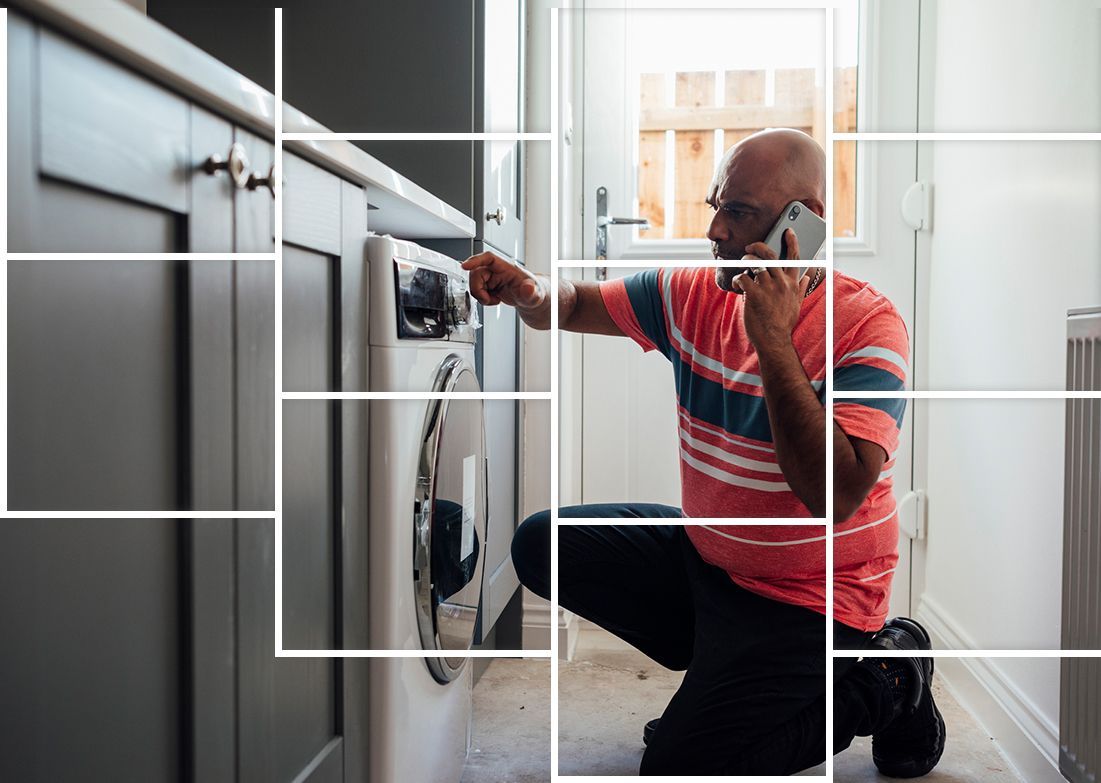A man is kneeling down in front of a washing machine while talking on a cell phone.