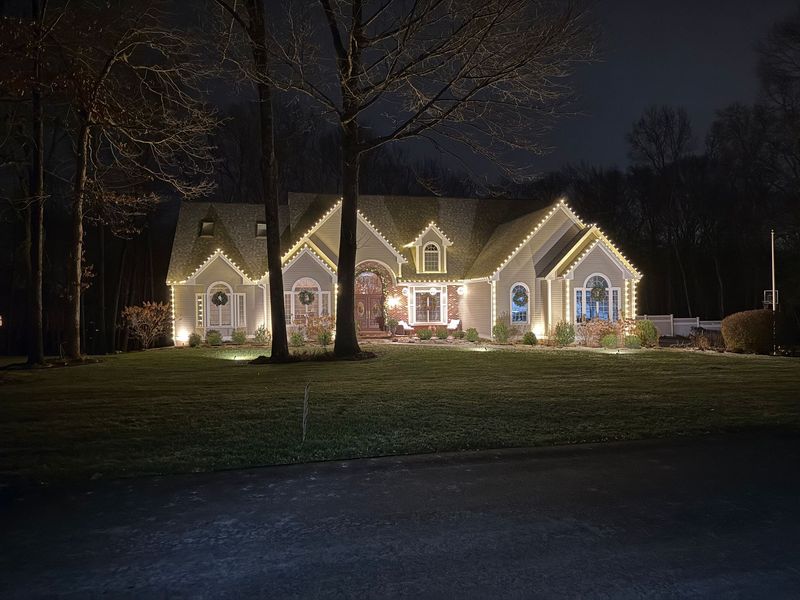 House at night, decorated with Christmas lights. Glowing lights trim roof, windows, and landscape.