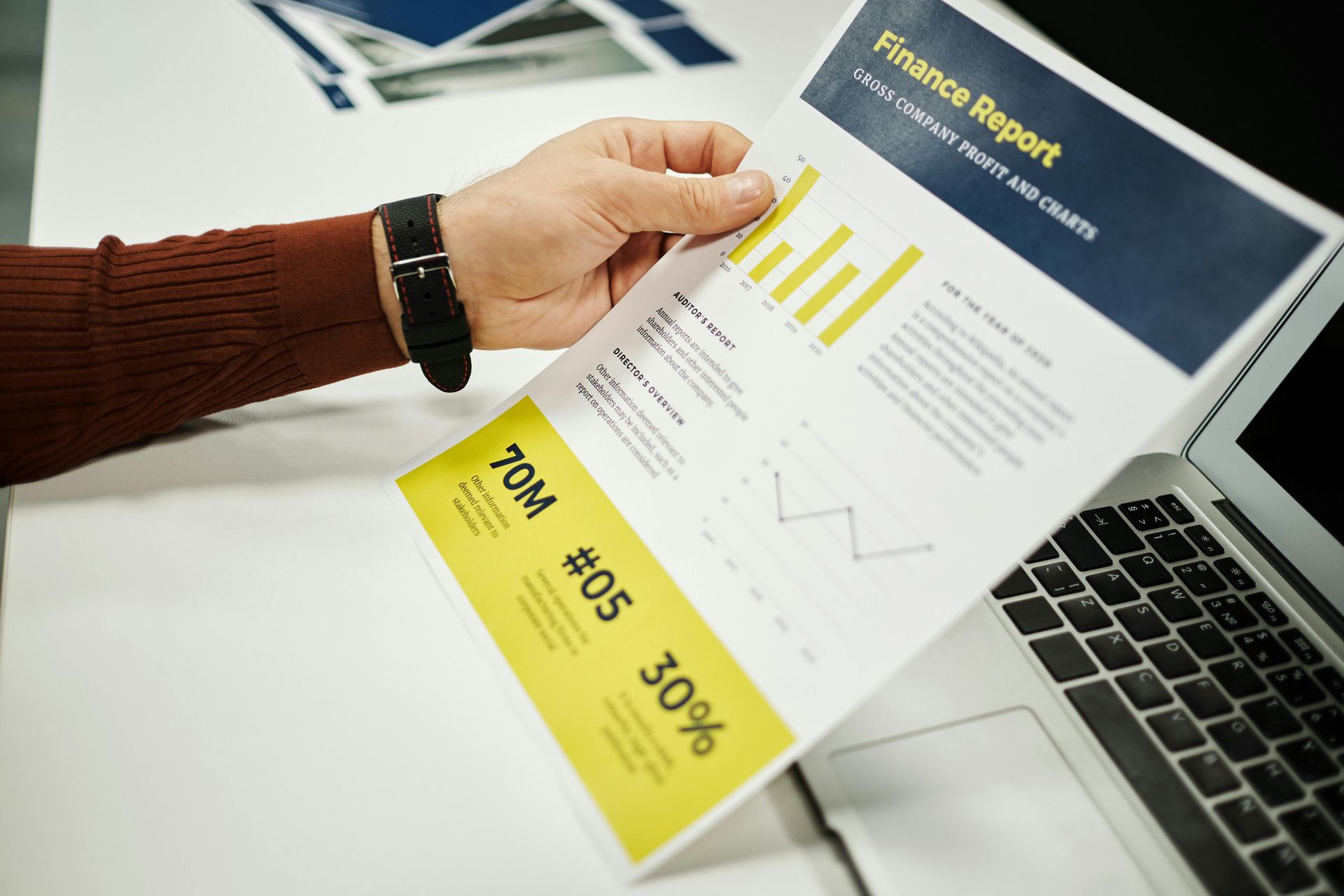 A person is holding a finance report in front of a laptop.