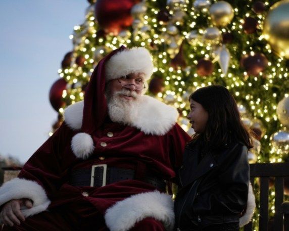 Santa Claus in red suit sits in a red chair, talking on the phone near a decorated Christmas tree.