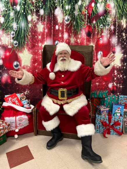 Santa Claus holding a wrapped red gift, pointing, smiling in a cozy room.