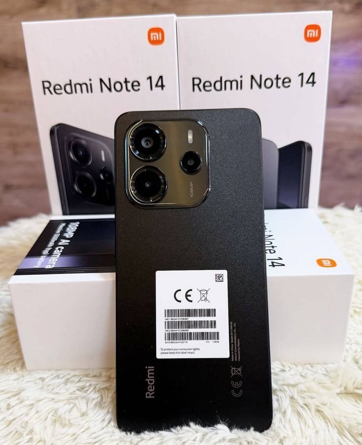 Teléfono Redmi Note 14 negro con cámara de triple lente, frente a dos cajas con el mismo modelo de teléfono.