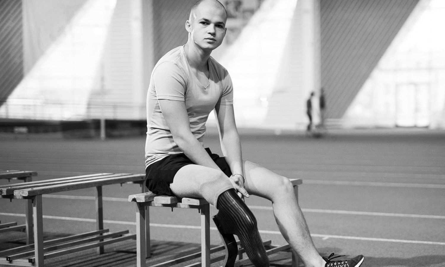 Black and white photo of a man wearing workout clothes with prosthetic legs sitting on a bench