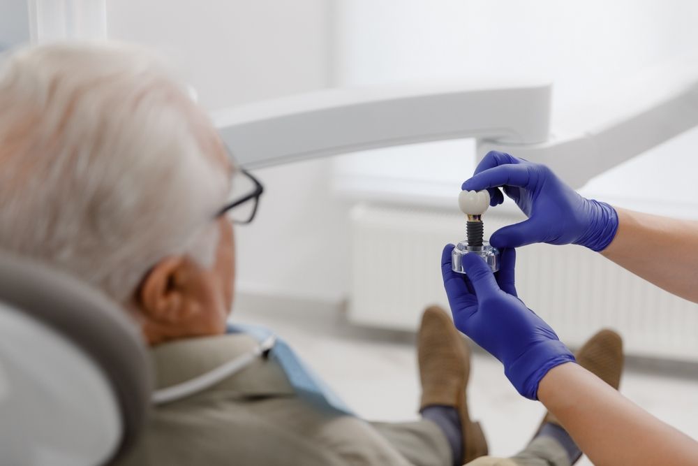 An elderly man is sitting in a dental chair while a dentist holds a dental implant.