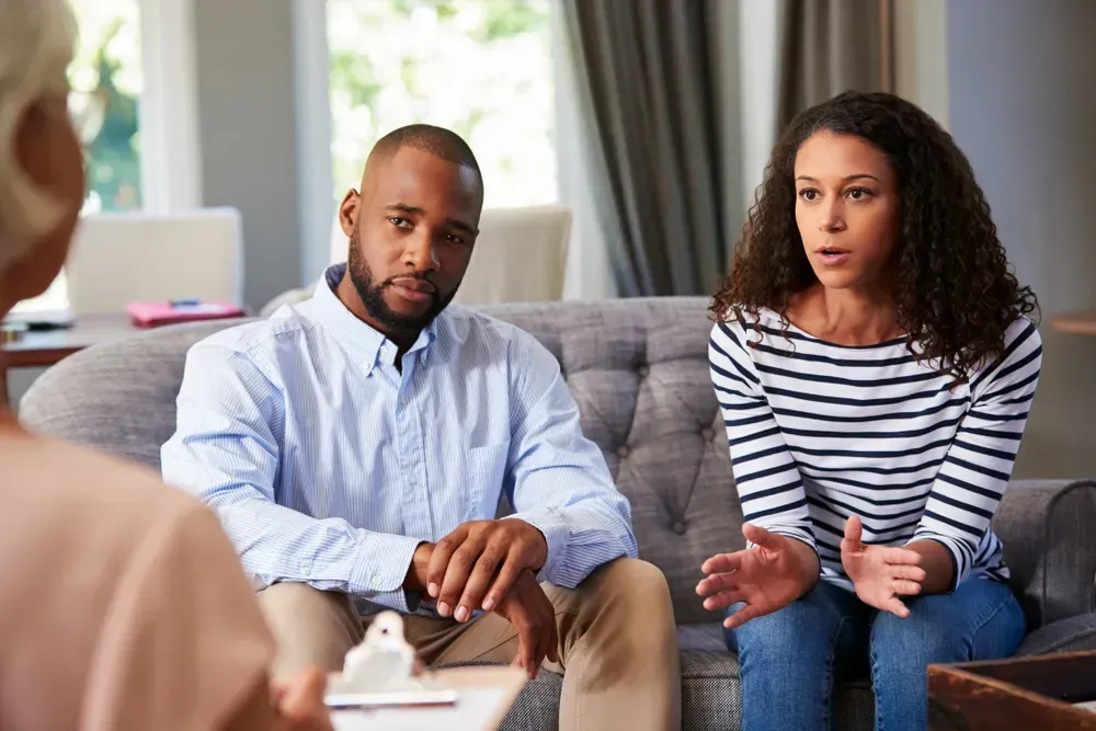 Couple on a couch talking to a person holding a clipboard, possibly a therapist's office.