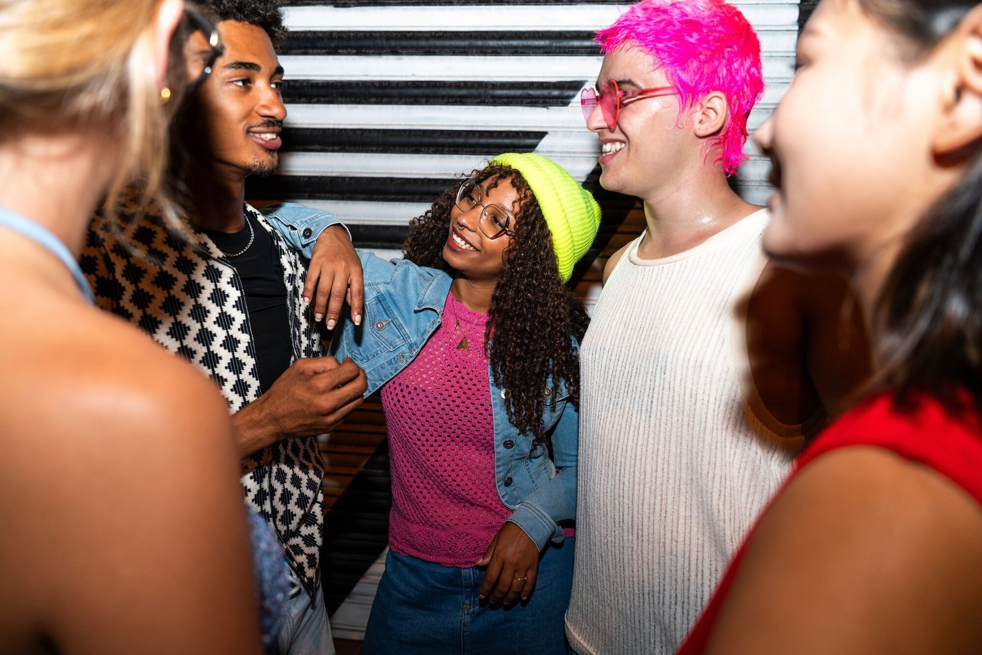 Group of friends chatting, smiling. Woman in neon green beanie, pink hair. Dark background, striped wall.
