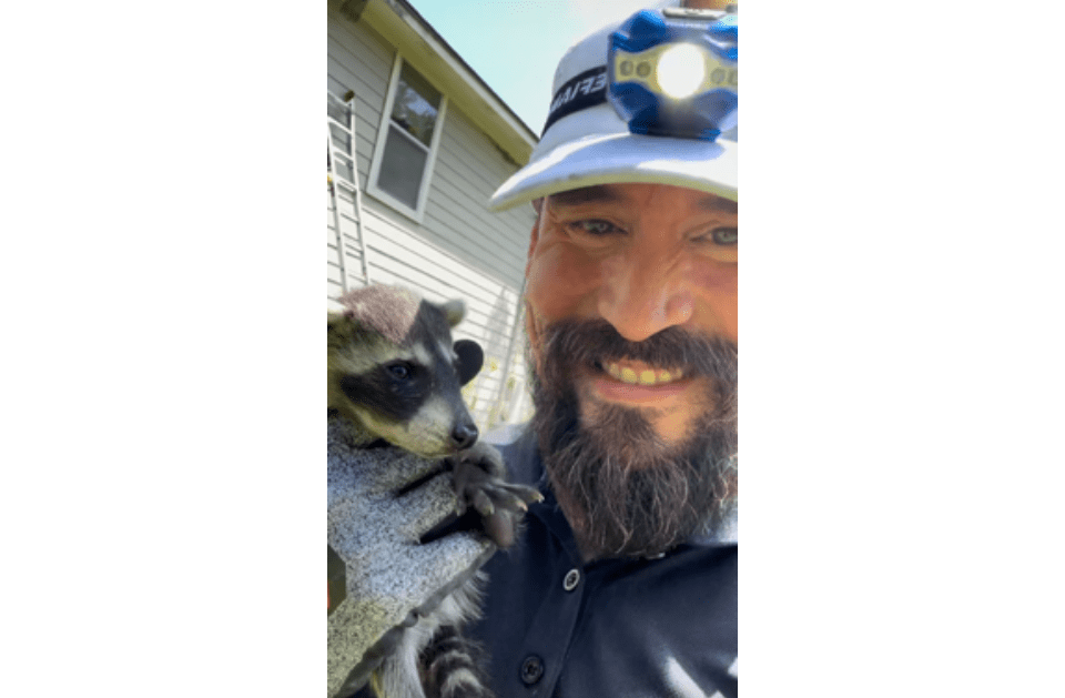 Man with dark beard holding a raccoon outdoors, both looking at the camera.