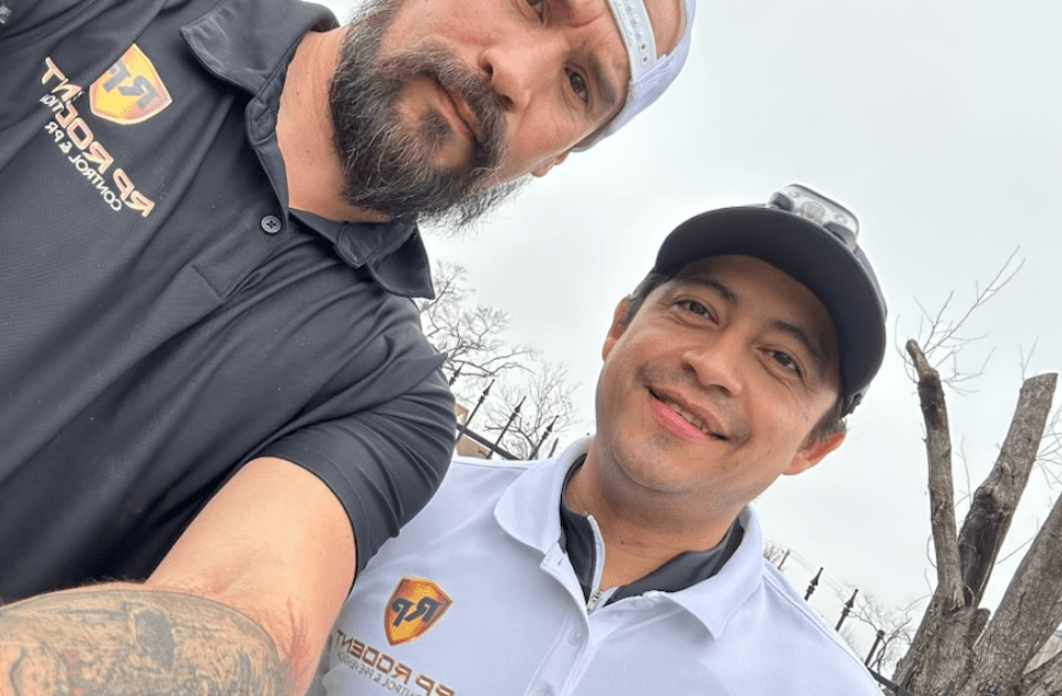 Two men taking a selfie outdoors; one with a beard and baseball cap, the other smiling in a white shirt.