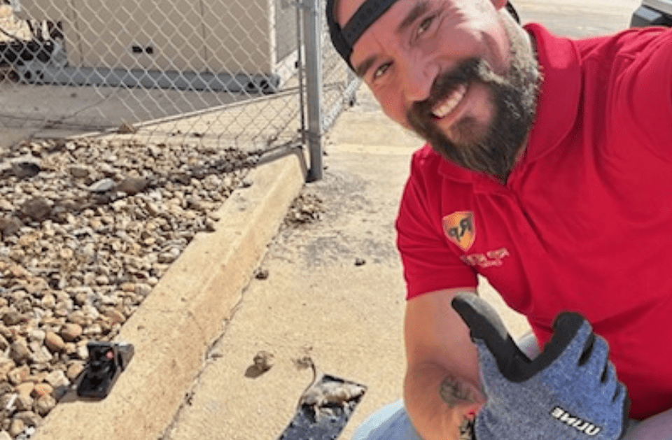 Man wearing a red shirt and black cap smiles while holding work glove, near a fence.