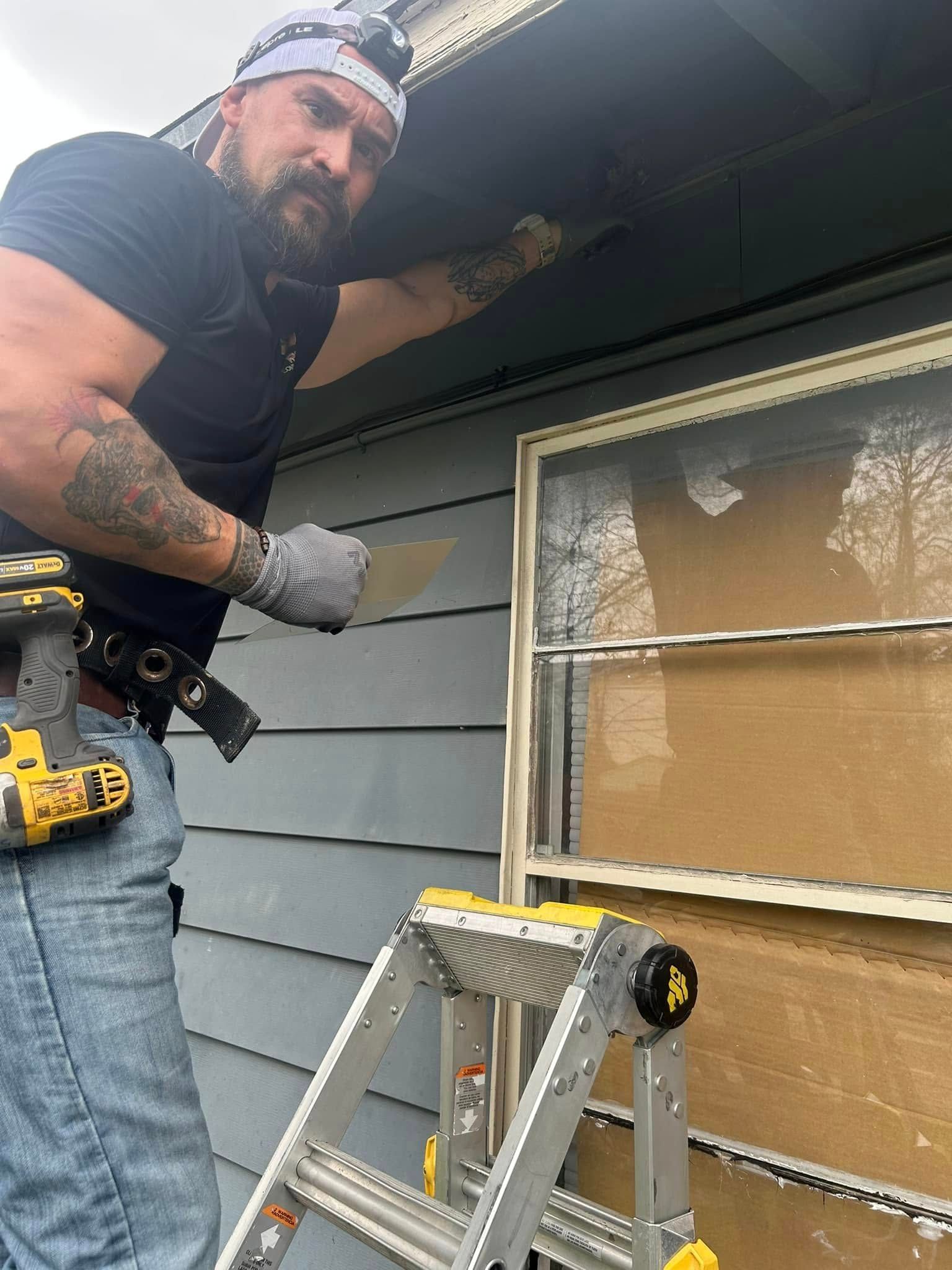 A person on a ladder repairs a house's siding. They wear a headlamp and hold a tool, with a serious expression.