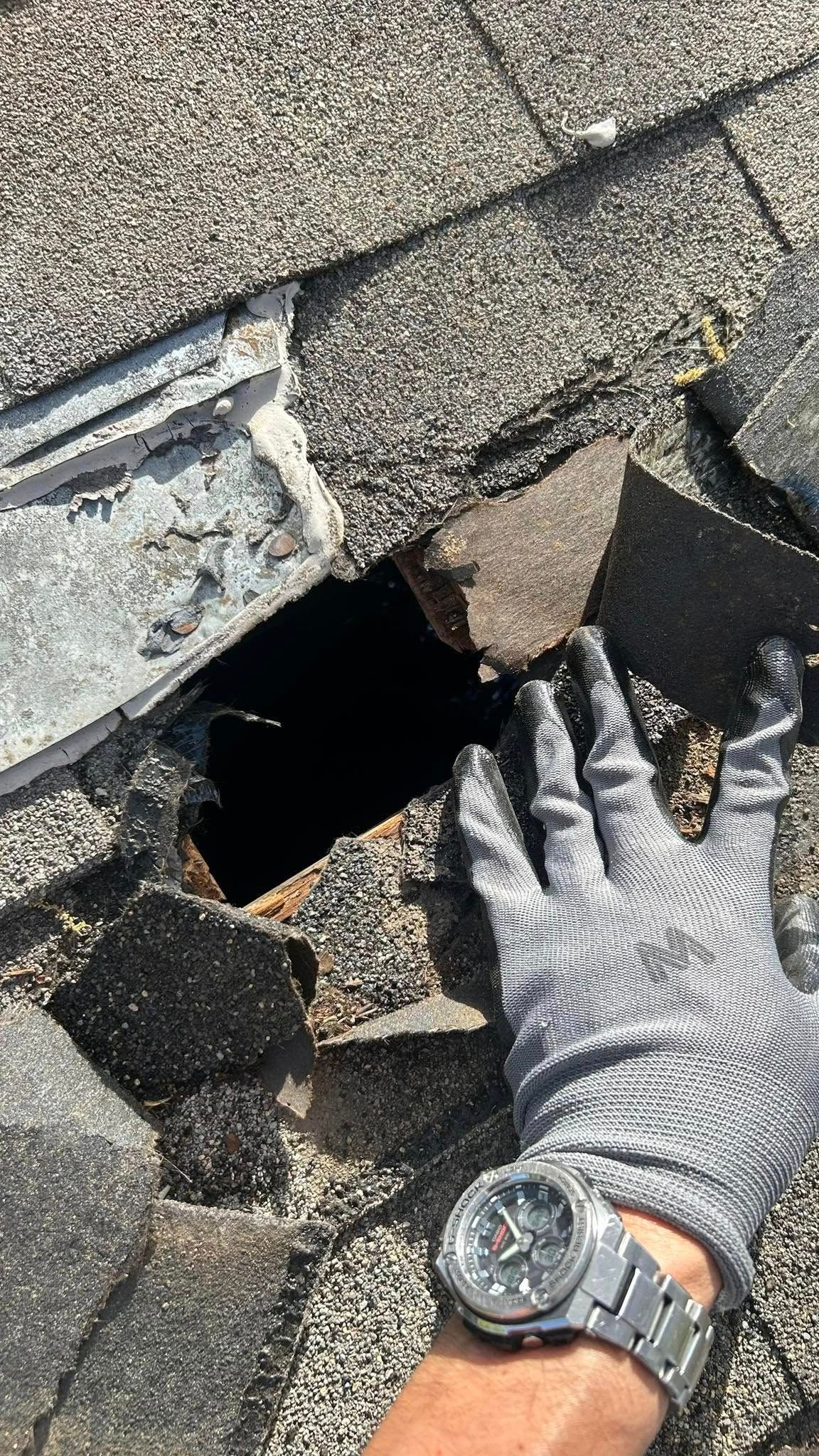 Gloved hand near hole in shingled roof, showing damage and potential water entry point.