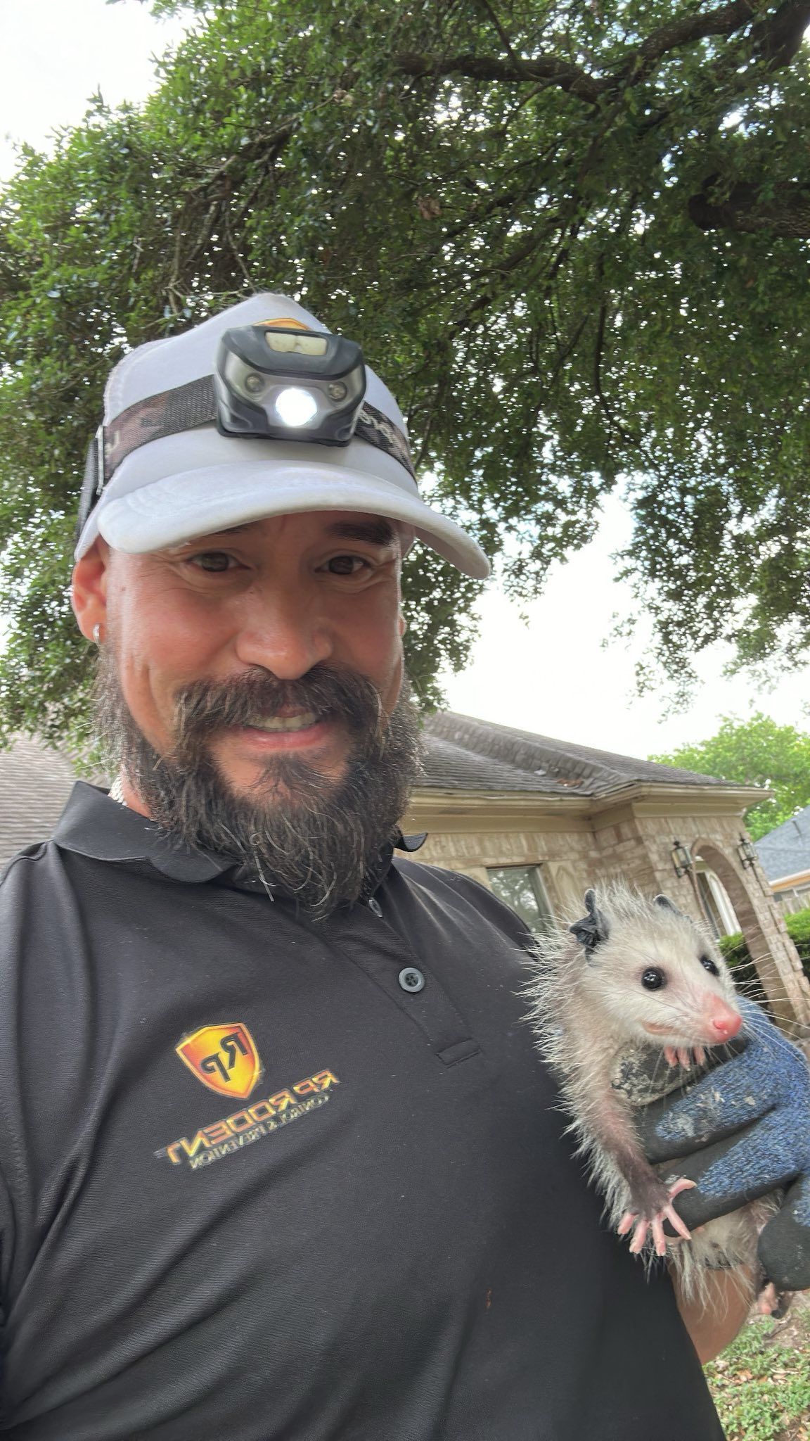 Man with headlamp holds an opossum in gloved hand; outdoors near a house.
