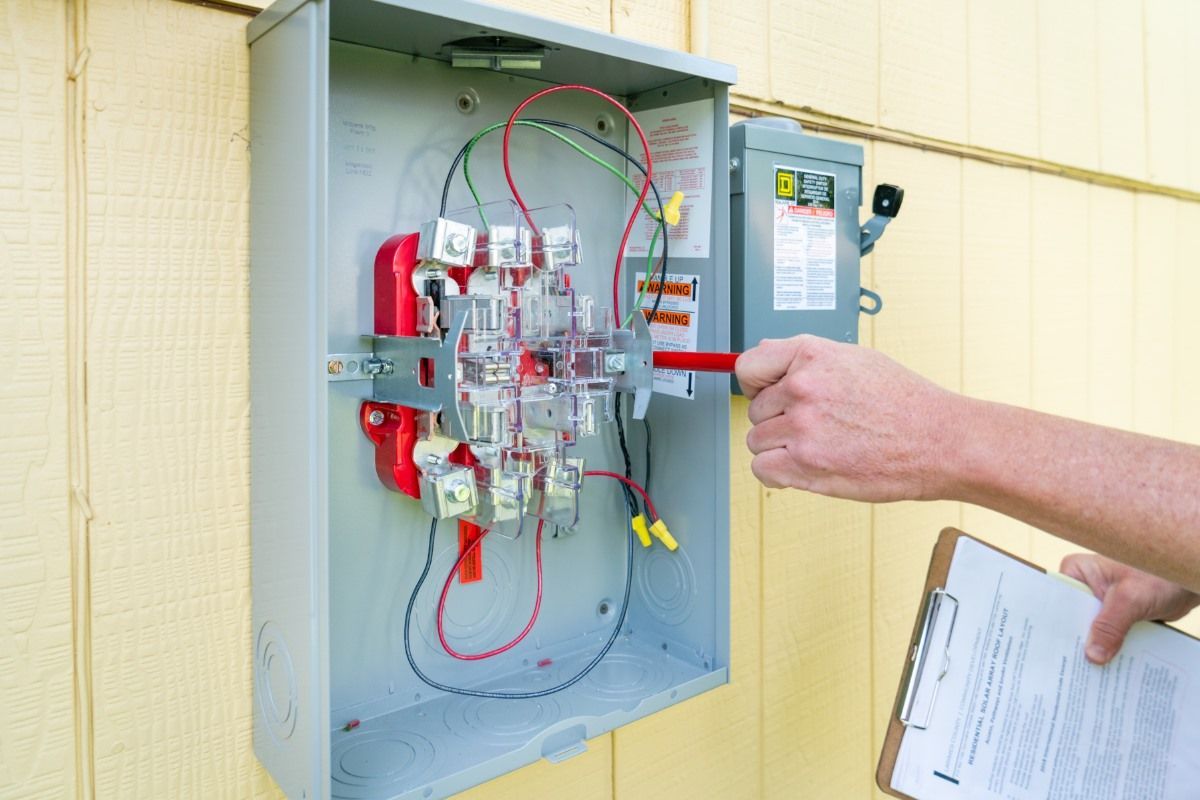 A person is working on an electrical box while holding a clipboard.