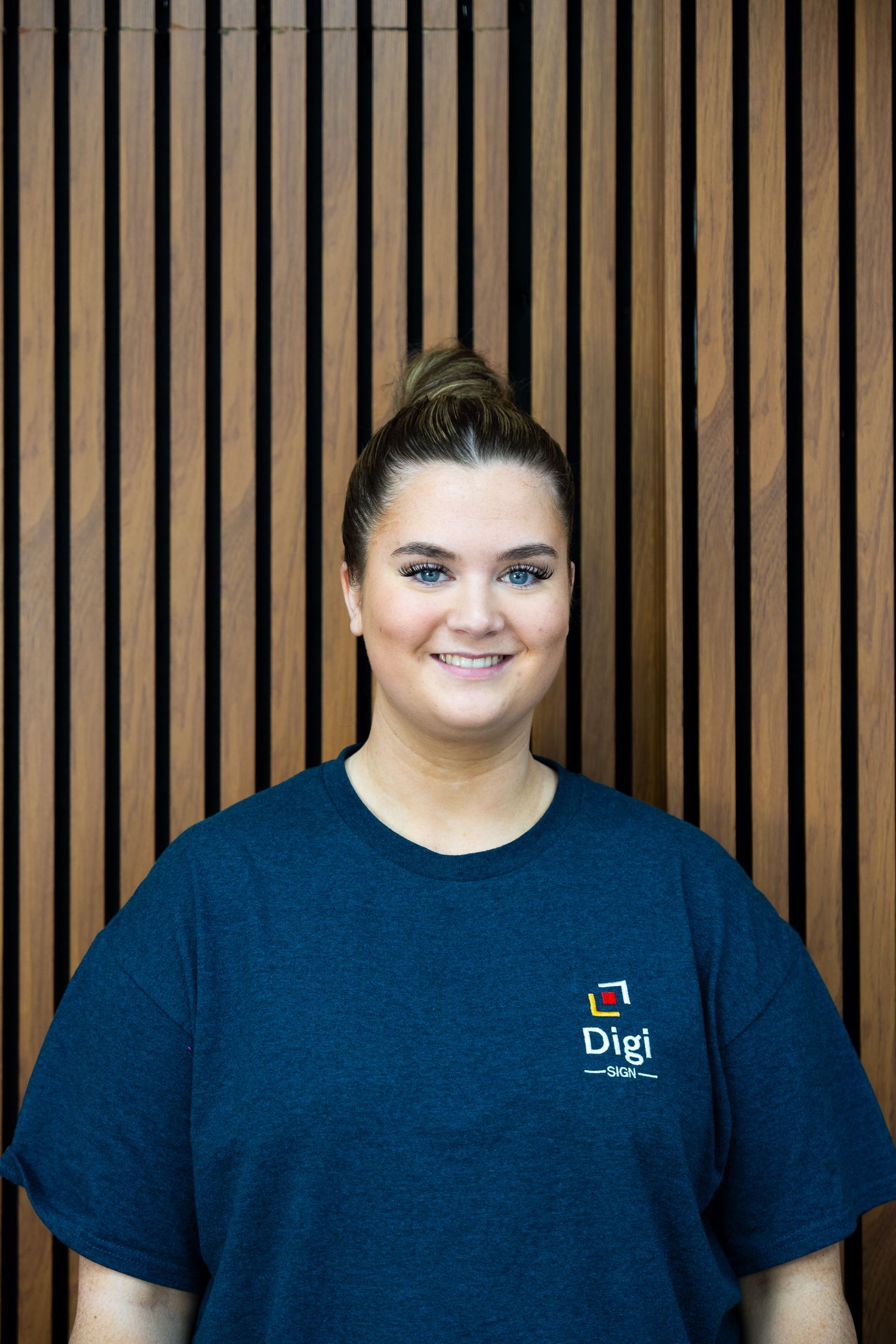 A woman wearing a blue t-shirt is standing in front of a wooden wall.