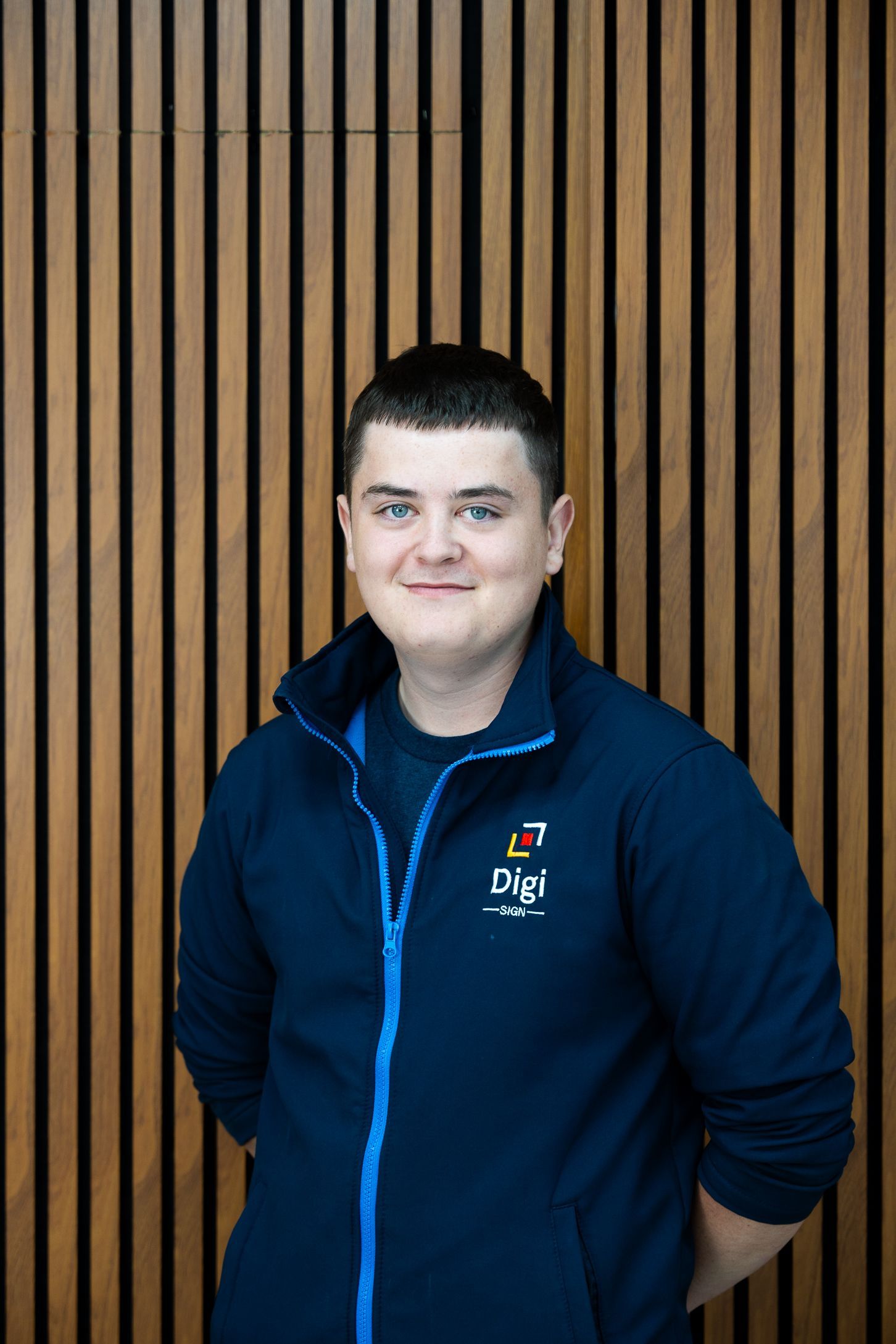 A young man in a blue jacket is standing in front of a wooden wall.