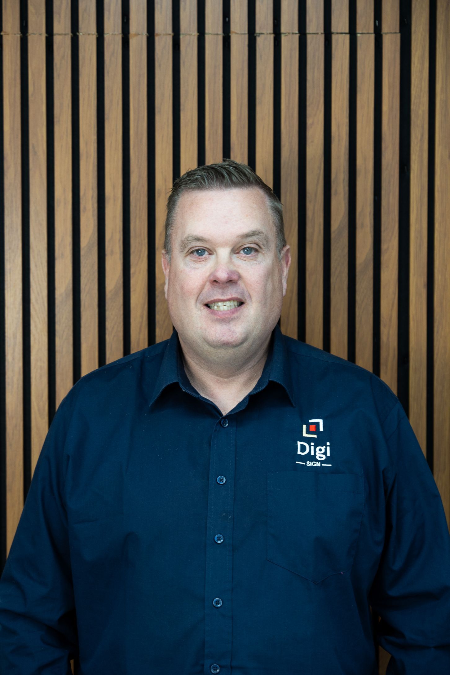 A man in a blue shirt is standing in front of a wooden wall.