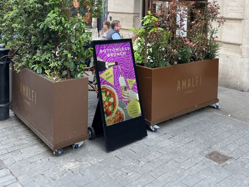 A sign with a picture of a pizza on it is sitting on the sidewalk in front of a restaurant.