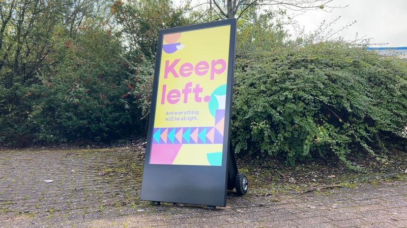 A large sign that says `` keep left '' is sitting in front of a bush.