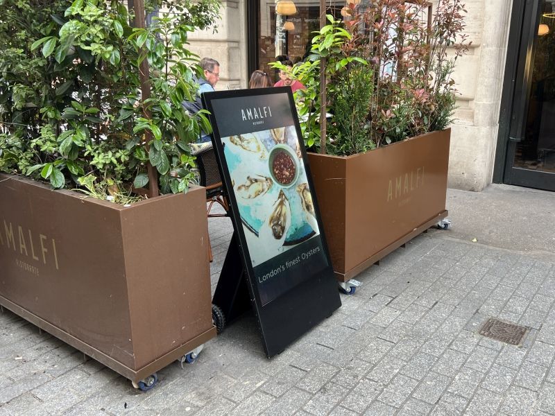 A digital sign is sitting on the sidewalk in front of a restaurant.