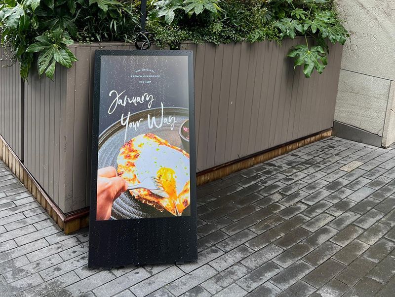 A sign with a picture of a pizza on it is sitting on a brick sidewalk.