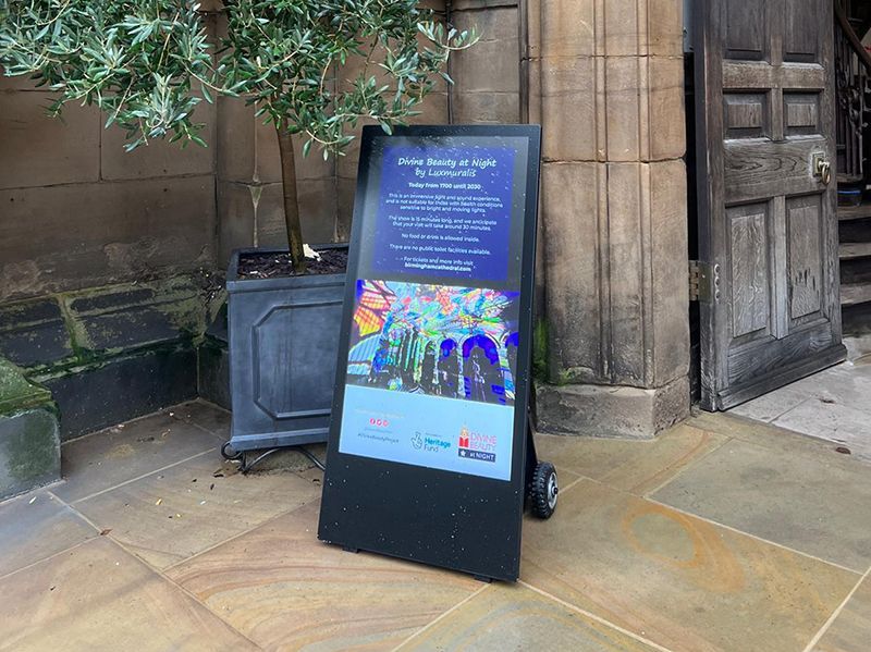 A digital sign is sitting on the sidewalk in front of a building.