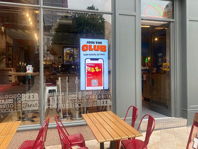 A restaurant with tables and chairs outside and a sign on the window that says `` join the club ''.